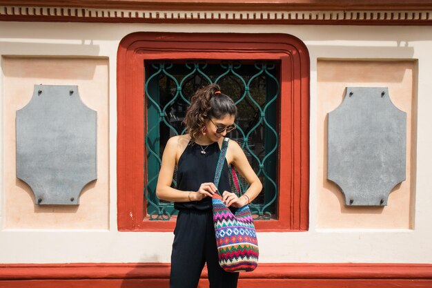 Young beautiful mexican woman shopping with sunglasses, black\
jumpsuit, and a colorful craft bag.