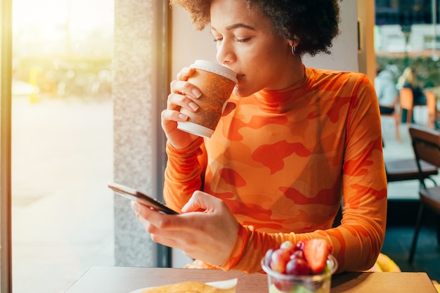 Caffè bevente della barra di seduta della giovane bella donna della corsa mista facendo uso del telefono