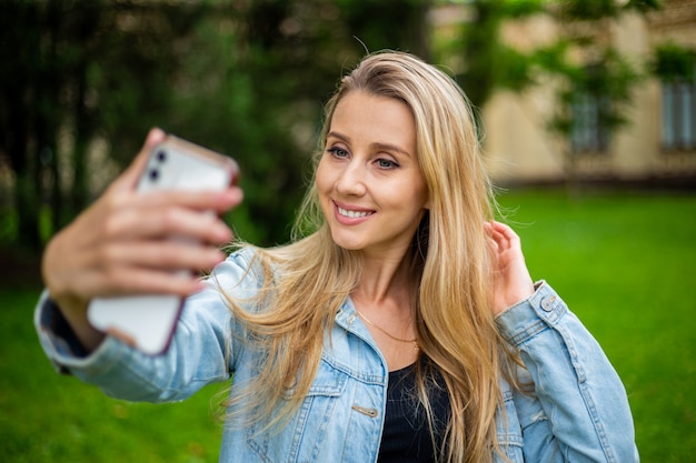 デニムジャケットを着た若い美しいモダンなファッショナブルな女の子が電話で自分撮りをします