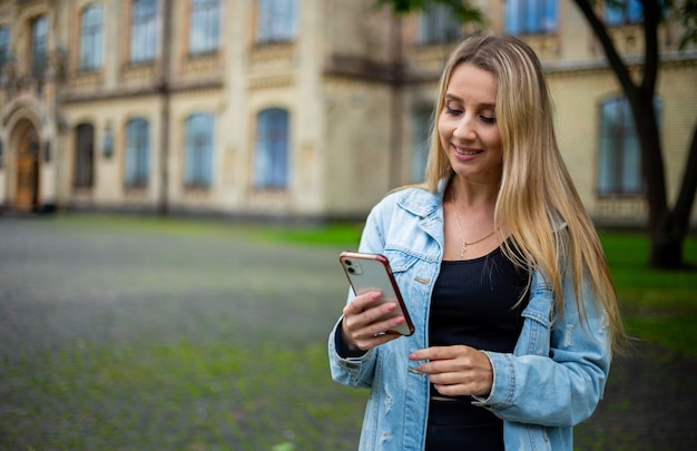 Young beautiful modern fashionable girl in a denim jacket on the street looking at the phone