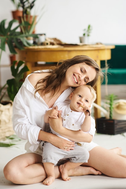 A young beautiful mom playing with her baby on the floor next to plants and green sofa