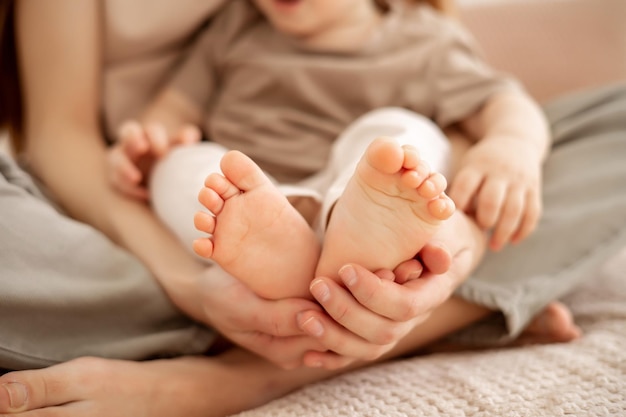 A young beautiful mom with a baby boy hugging on the bed at home in the bedroom mom holding the baby's small legs in her hands baby feet closeup
