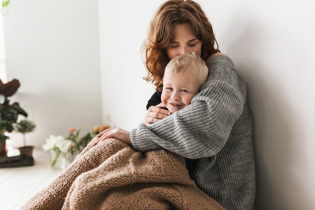 Young beautiful mom with red hair in knitted sweater sitting on floor, kissing her little son