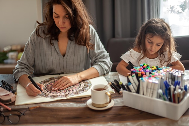 Young beautiful mother and cute daughter draw a mandala pattern together artistic markers and colore...