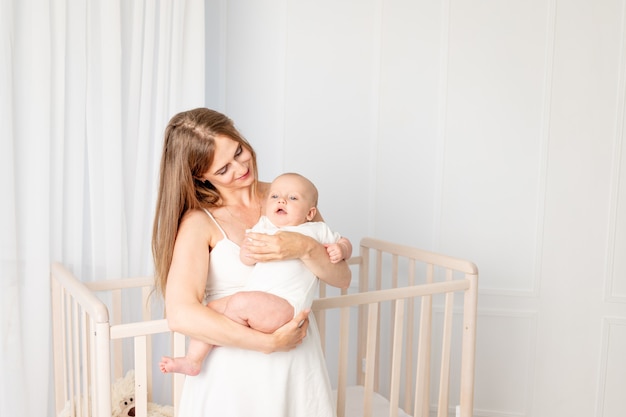 Young beautiful mother holding her daughter 6 months, hugging her in the nursery standing by the crib