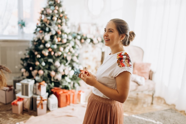 Young beautiful mother holds New Year's decorations standing in the full of light cozy room next to the New Year's tree .