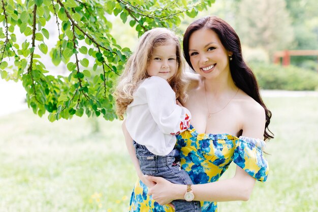 Young beautiful mother hugs her cute daughter holds her in her arms standing in the park near a tree