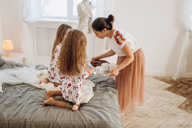 La giovane bella madre sta portando il cacao con i marshmallow e i biscotti alle sue figlie sedute sul letto nella stanza piena di luce e accogliente.