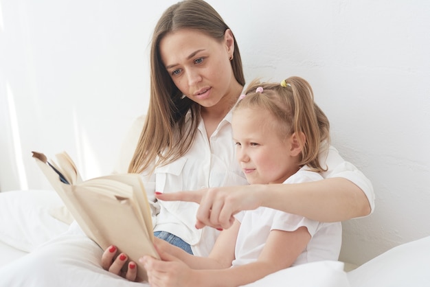 La giovane bella madre sta leggendo un libro a sua figlia. famiglia felice, madre e figlia che leggono un libro mentre sono sdraiati a letto, mamma tata sorridente che racconta storie divertenti a una ragazza prescolare carina.