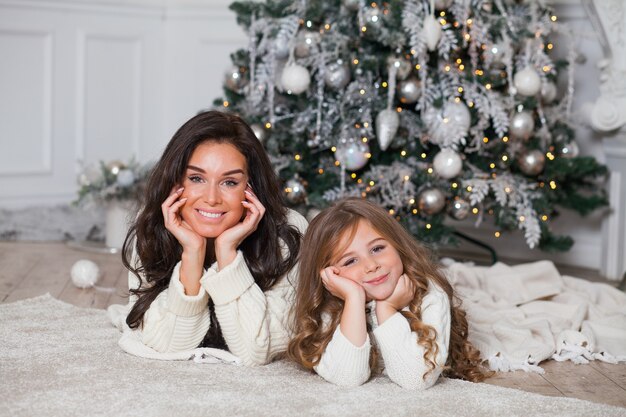 Young beautiful mother and little cute daughter in white cozy sweaters and blanket have fun and hug under a decorated Christmas tree. Festive home classic interior.