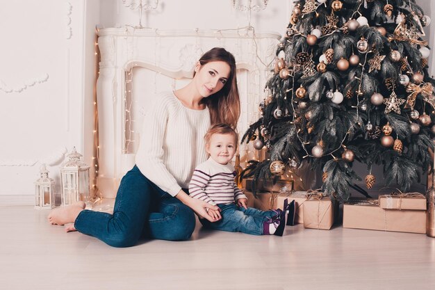 Young beautiful mother sitting with baby girl under in room with Christmas tree