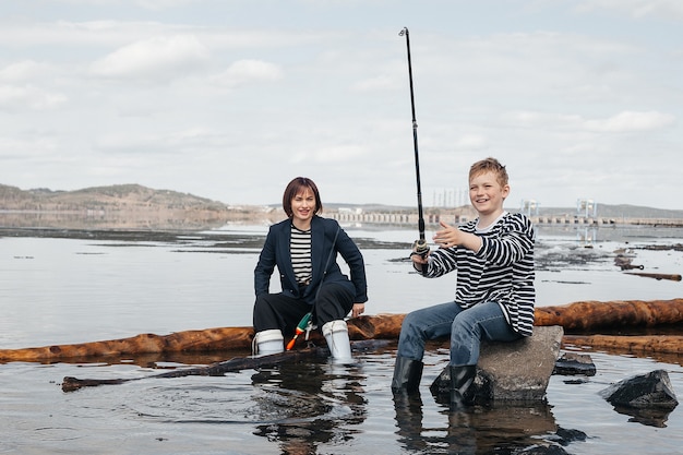 Young, beautiful mother and son have fun fishing on the lake. Mom and son in striped vests on the river bank with a fishing rod.