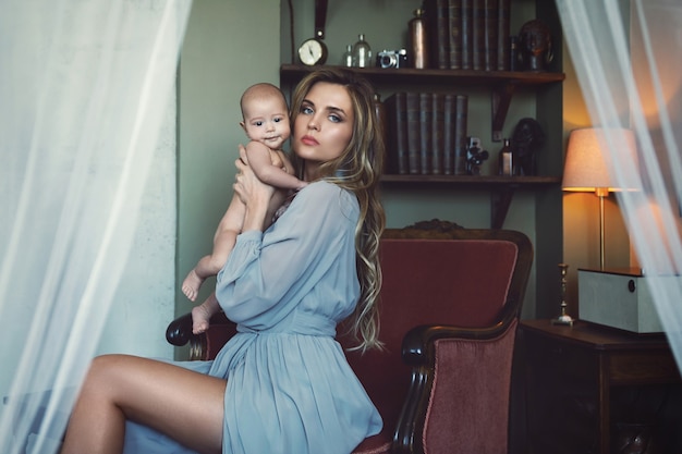 Young beautiful mother wearing luxury grey dress sitting in the armchair and holding her baby on hands