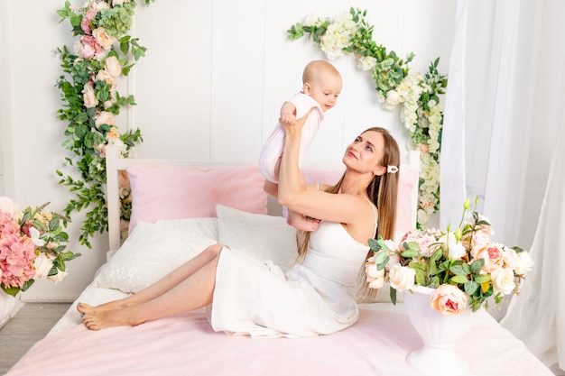 A young beautiful mother with a 6-month-old daughter in her arms sits on a white bed in flowers and hugs her, a place for text