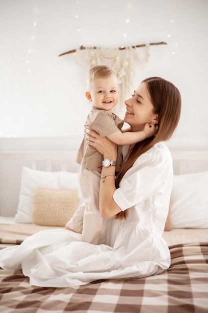 A young beautiful mother with a baby boy hugging on the bed at home in the bedroom mom's care and love children and parents