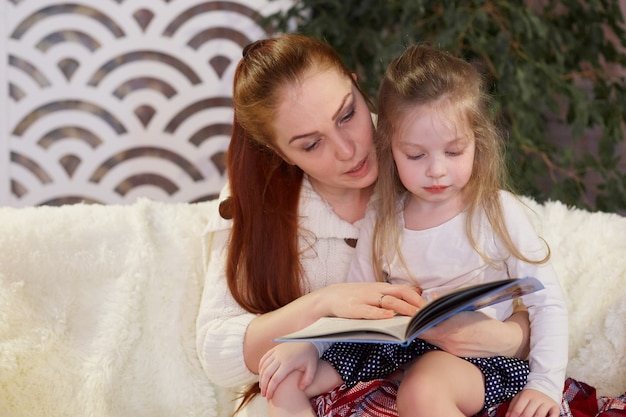 Young beautiful mother with her little daughter reading a book sitting on the couch.