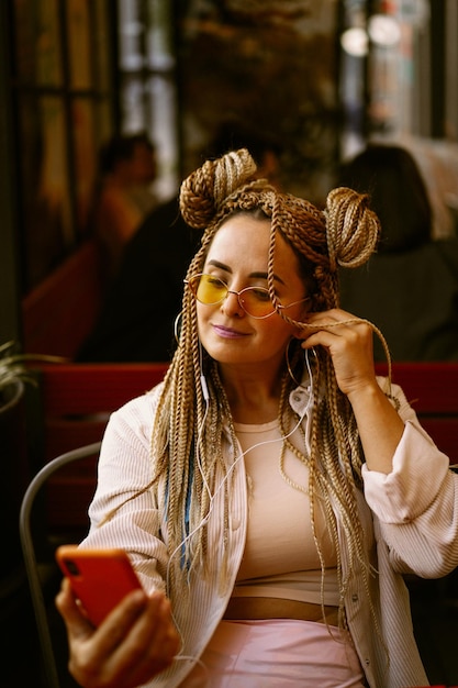 Young beautiful multiracial woman with blonde afro hairstyle braids zizi on the street cafe with headphones. yellow sunglasses, bright makeup, hippie style. video call, work outside the home