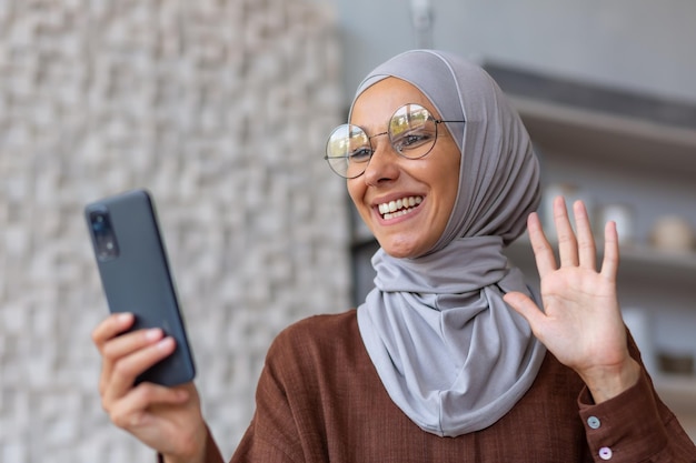 ヒジャブと眼鏡を着た若い美しいイスラム教徒の女性が、家で手を振って電話からビデオ通話で話している