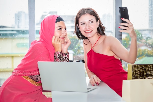 Young beautiful Muslim woman selfie with Caucasian friendships sitting near shopping bags and tablet enjoying in shopping at coffee shop. 