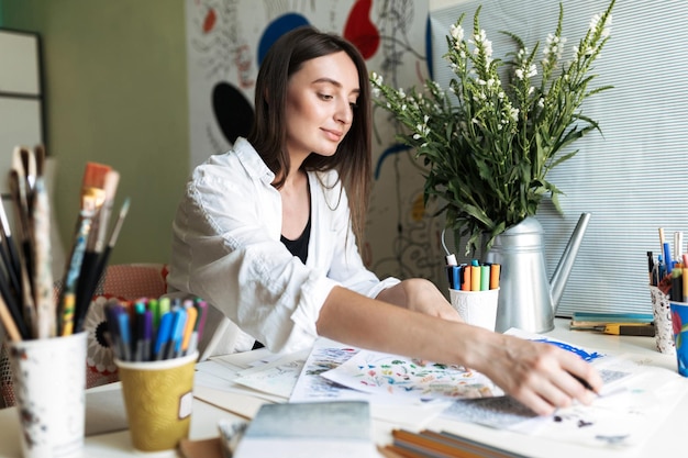 Young beautiful painter sitting at the desk with pictures dreamily working at cozy home