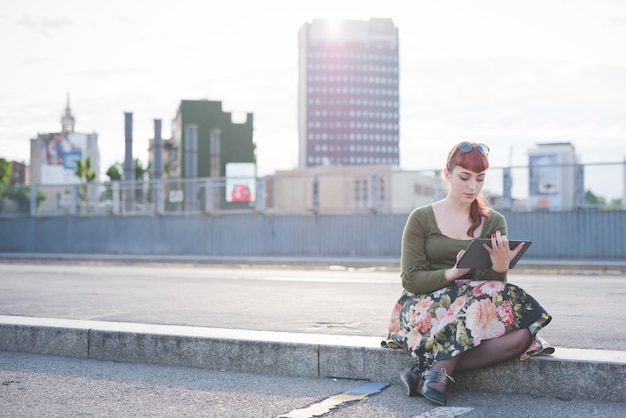 Young beautiful pin up redhead woman sitting on a sidewalk in the city back light