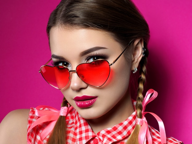 Young beautiful playful woman looks over her heart shaped red glasses. Valentines day, love or theme party concept. Smokey eyes and red lips makeup. 