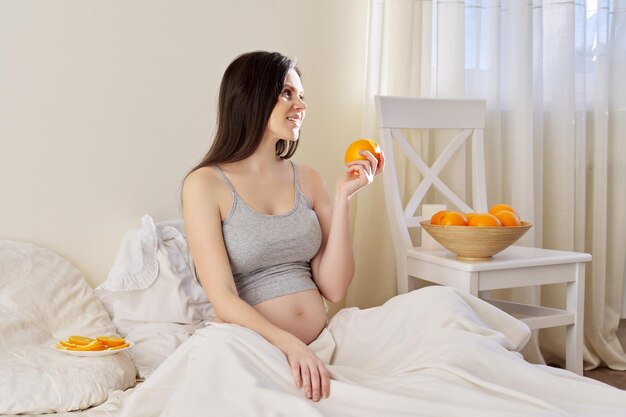 집에서 침대에 앉아 오렌지를 먹는 젊은 아름 다운 임신한 여자. 건강식, 천연비타민C