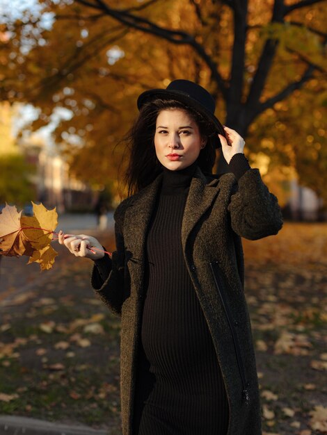 Giovane bella donna incinta con un cappello con i capelli scuri in un vestito attillato nero e una posa del cappotto