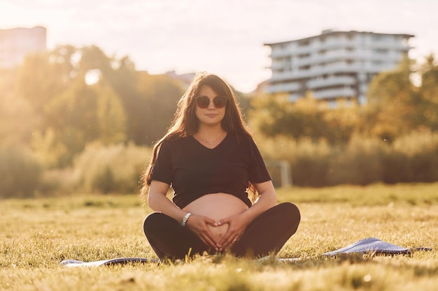 Young beautiful pregnant woman that is in sunglasses is doing\
yoga on the mat outdoors on the field