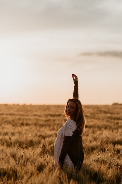 Foto giovane bella donna incinta in vestito bianco che cammina nel campo di grano arancione in una soleggiata giornata estiva. senti la libertà con il braccio teso al cielo. aspettativa miracolosa. tramonto sull'isolamento. abbagliamento del sole.