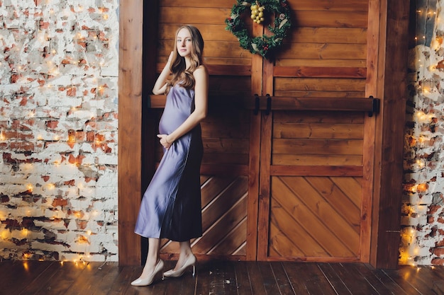 Молодая красивая беременная женщина со светлыми волосами и нежным макияжем в модном платье позирует рядом в студии