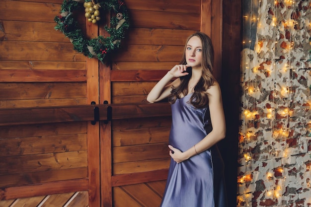 Young beautiful pregnant woman with blond hair and gentle makeup in fashionable dress posing near at the studio