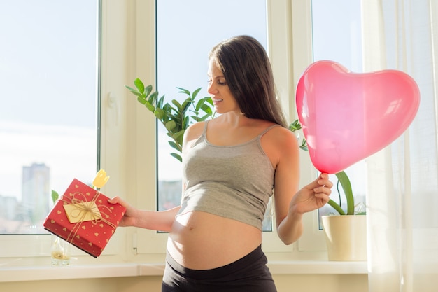 Giovane bella donna incinta con scatola regalo e palloncino cuore