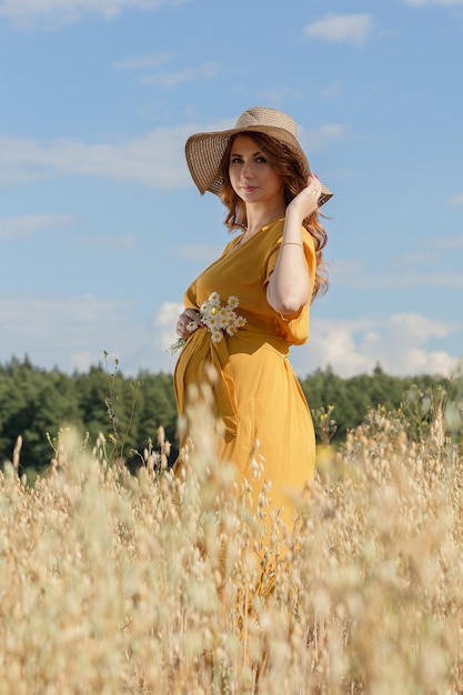 A young beautiful pregnant woman in a yellow dress and hat walks through a wheat orange field on a sunny summer day