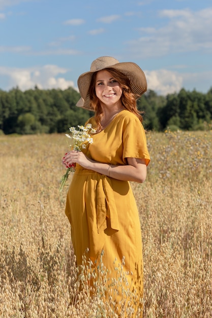 A young beautiful pregnant woman in a yellow dress and hat walks through a wheat orange field on a sunny summer day