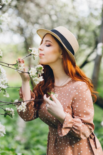 Young beautiful red-haired girl walks in a spring blooming apple orchard