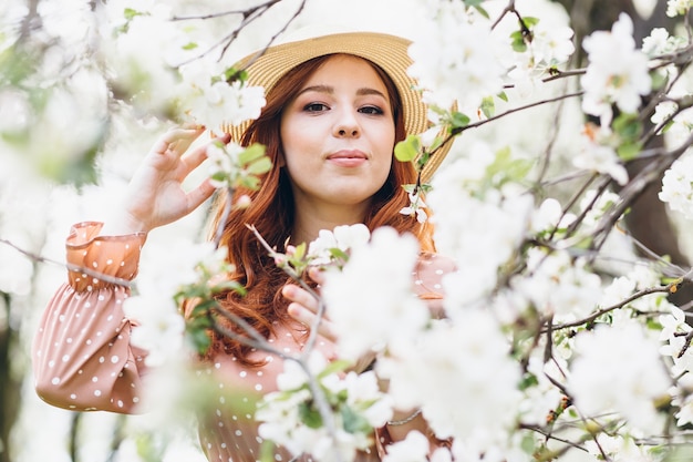 Young beautiful red-haired girl walks in a spring blooming apple orchard