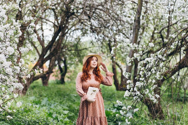 Young beautiful red-haired girl walks in a spring blooming apple orchard