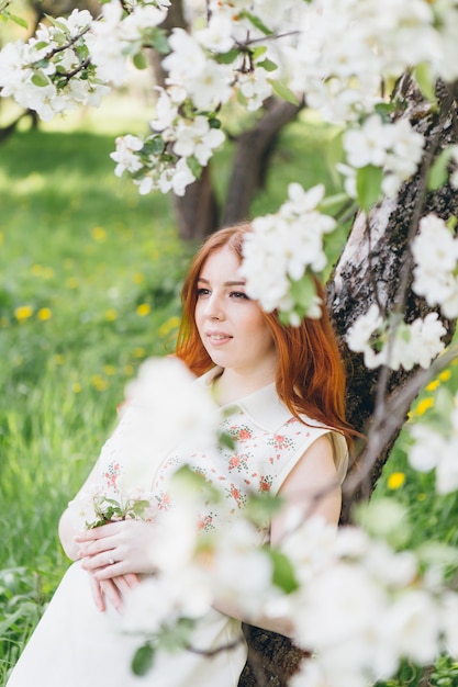 Young beautiful red-haired girl walks in a spring blooming apple orchard