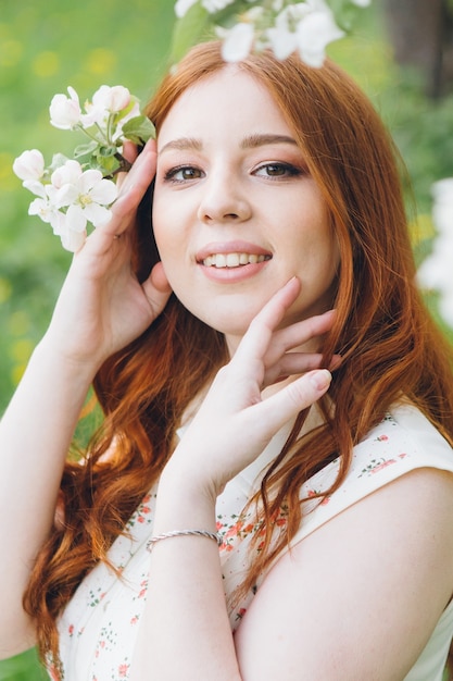 Young beautiful red-haired girl walks in a spring blooming apple orchard