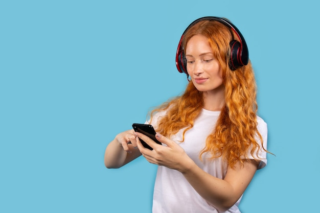 A young beautiful red-haired girl in a white T-shirt chooses music on the phone and enjoys it in big headphones. Photo on blue wall with empty side space. 