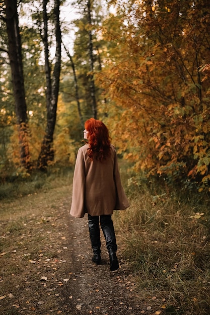 Young beautiful red headed woman in beige coat walking in autumn forest