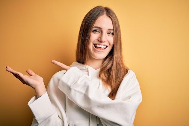 孤立した黄色の背景の上にスエット シャツを着ている若い美しい赤毛のスポーティな女性は驚き、手で提示し、指で指しながらカメラに笑顔