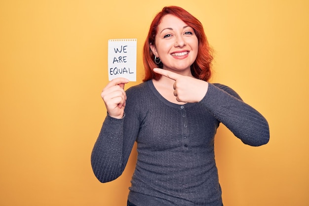 Young beautiful redhead woman asking for equality holding paper with we are equal message smiling happy pointing with hand and finger