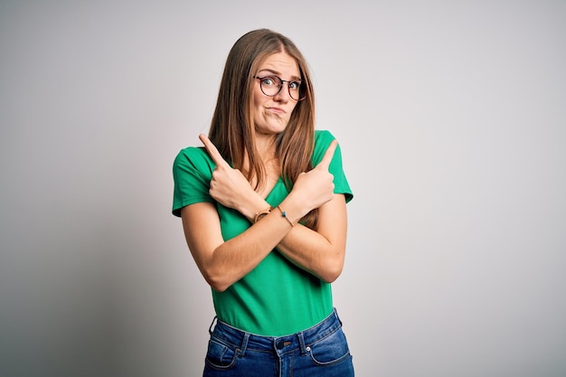 Young beautiful redhead woman wearing casual green tshirt and glasses over white background Pointing to both sides with fingers different direction disagree