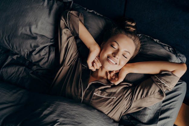 Молодая красивая расслабленная брюнетка женщина в атласной пижаме растягивается в постели