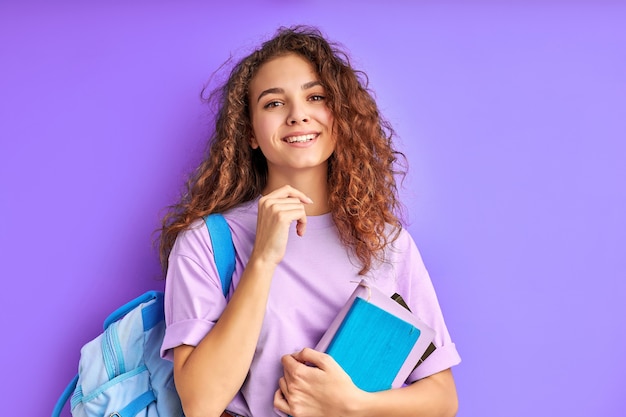 Young beautiful school girl with curly hair keen on studying, preparing for school or college, isolated purple space