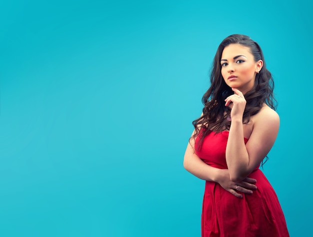 Young beautiful and serious girl in a red dress, with makeup on a blue wall