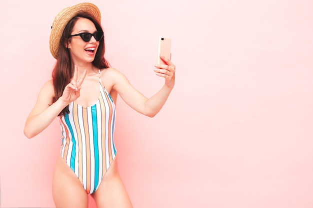 Foto giovane bella donna sorridente sexy dei pantaloni a vita bassa nella posa vicino alla parete azzurra in studio. modello alla moda in costume da bagno colorato estivo. modello positivo che prende selfie. felice e allegro con il cappello