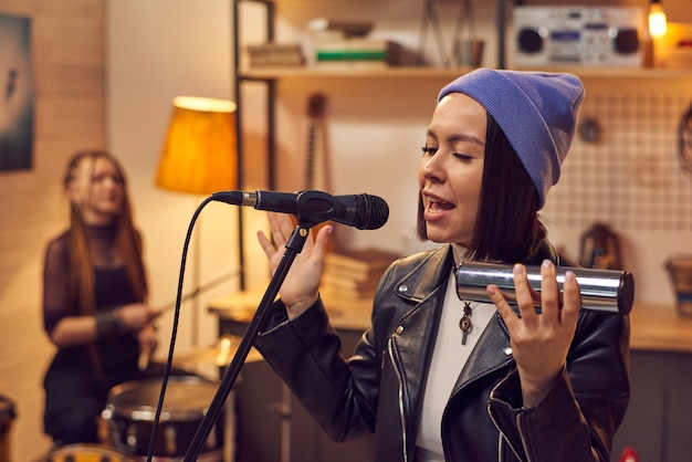 Young beautiful singer in stylish hat playing musical instrument and singing in microphone in music studio